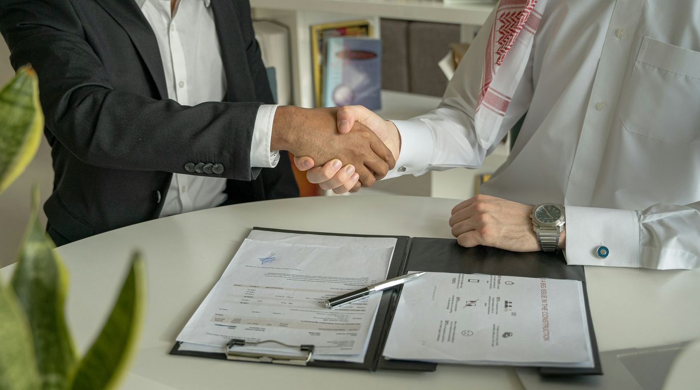 Two businessmen shaking hands across a table, with a contract and a pen in front of them